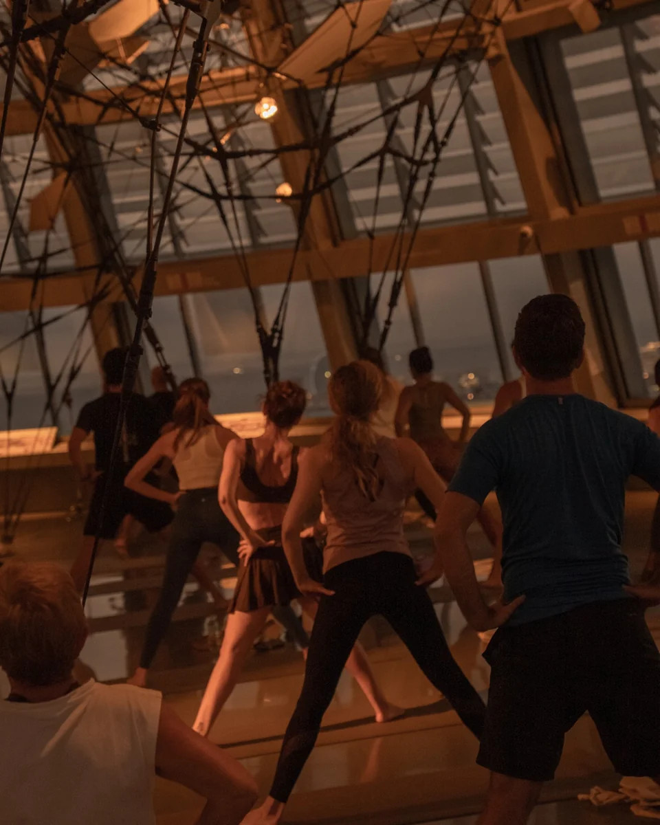 Grupo de participantes realizando posturas de yoga de pie frente a los ventanales del Mirador Torre Glòries en Barcelona.